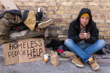Homeless Man Sitting On The Street, Near The Wall And Cart With His Stuff. Holding Cup Of Coffee And Looking At Camera.