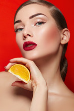 Beautiful Young Girl In The Studio On An Orange Background Holds In Her Hand A Slice Of Orange. Smooth Skin And Hair. Styling, Shiny Hair. Fruits, Health, Diet, Fashion, Beauty. Makeup - Red Lips.