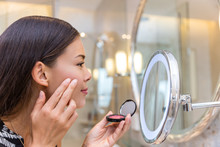 Asian Young Woman Putting Blush Cream Using Fingers While Looking In Makeup Mirror At Home In Bathroom. Morning Routine Of Girl Applying Make-up Before Going To Work. Face Skincare.