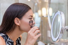 Woman Using Eyelash Curler Curling Eyelashes Before Mascara In Lighted Round Makeup Mirror From Luxury Hotel Or Home Bathroom. Beautiful Asian Girl Getting Ready In The Morning Looking At Reflection.