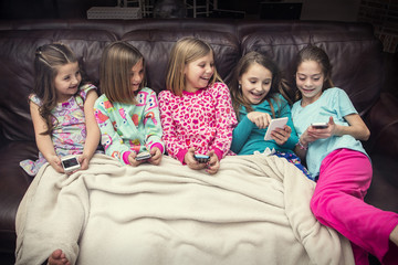 Group of cute, happy little girls playing with their smart phones together while sitting on the couch at a sleepover. Five girls all connecting to social media and reacting together to funny content