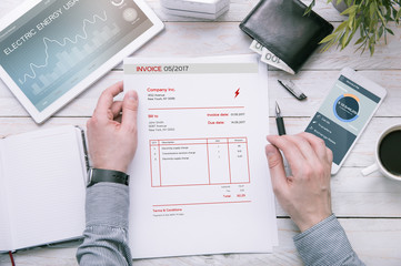 Man holds invoice of electric power usage over desk with tablet and smartphone with applications made in graphic program.