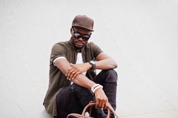 Portrait of sitting stylish african american man wear on sunglasses and cap with handbag outdoor. Street fashion black man.