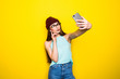 Trendy beautiful cool hipster girl taking selfie with mobile phone against a yellow wall.