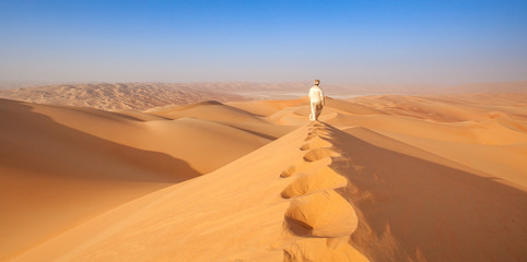 Wall Mural - arab man in Kandoura walking over a Dune in the arabian Desert