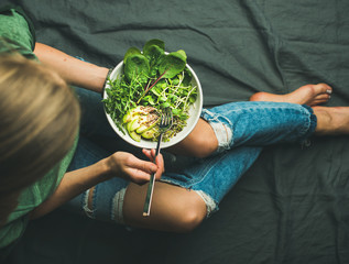 Wall Mural - Green vegan breakfast meal in bowl with spinach, arugula, avocado, seeds and sprouts. Girl in jeans holding fork with knees and hands visible, top view. Clean eating, detox, vegetarian food concept