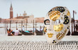 Traditional Venetian Mask with moored gondolas, romantic couple and San Giorgio Maggiore church in the blurred background. Hot summer holiday feeling in beautiful Venice. Authentic travel concept.