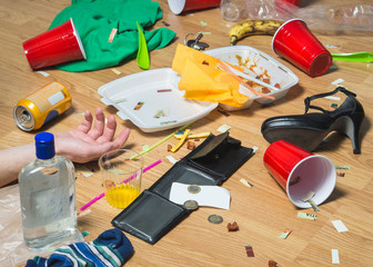 Man passed out on the floor after wild partying, hand visible. Terrible hangover and mess in the apartment. Trash, food leftovers, clothes, high heels and bottles everywhere. Regret and remorse.