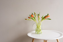 Close Up Of Orange Tulips In Glass Jug On Round White Table Against Neutral Background