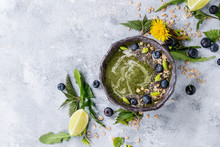 Spring Green Nettle And Dandelion Smoothie Bowl Served With Lime, Yellow Flowers, Young Leaves, Oat Flakes, Chia Seeds, Blueberries, Cream Over Gray Blue Texture Background. Top View, Space