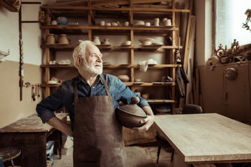 Front view of proud senior potter in apron standing with ceramic bowl at workshop