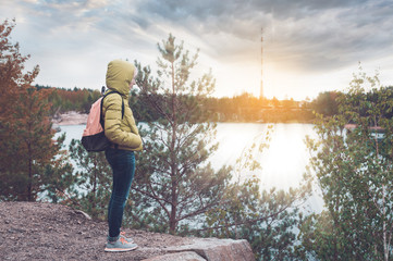 A female traveler with backpack enjoying the view of the lake on peak of rock among the pine forest. Tourist traveler.