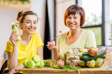 Young and older women sitting with healthy food and fresh drinks after the sports training indoors on the window background