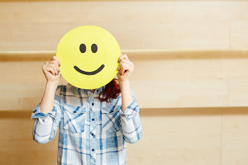 Unrecognizable young woman in checked shirt hiding her face under smiling mask while standing against wooden wall, waist-up portrait