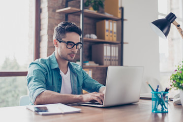young handsome minded man in jeans shirt and spectacles sitting at the table and typink a letter to 