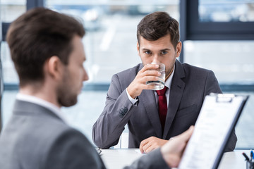 Nervous young man drinking water from glass during job interview, business concept