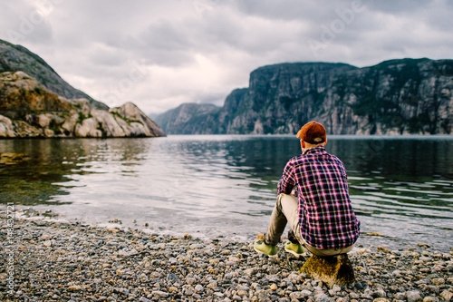 Lonely traveler tourist man resting on shore of fjord in Norway with far view of water and mountains valley at horizon in summer cloudy day Poster