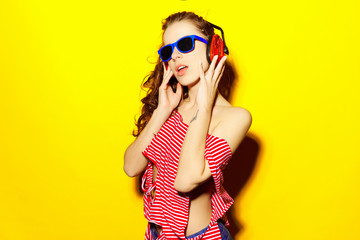 Beautiful young sexy girl dj in blue sunglasses and red striped T-shirt laughing and having fun and listening to music in red headphones on a yellow background