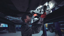 African Mechanic Working On The Underside Of A Car Elevated On A Hoist Shining A Bright Light Onto The Chassis