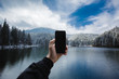 © illia_hnuskin - A man using smartphone takes pictures of the beauty of the forest mountains on the lakeshore. A blue sky and clouds are in the background