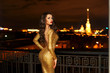 Sexy fashionable beautiful young woman in long golden shining evening dress standing at balcony over view on night city lights in bokeh. Pretty lady with long curly hair and red lips