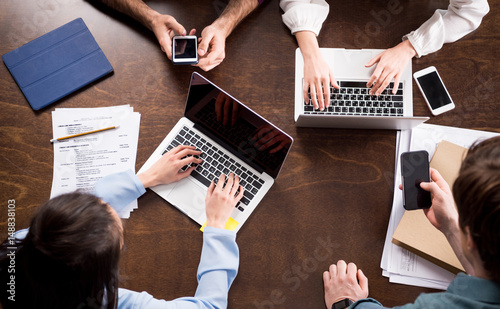 Fotomural Overhead view of young businesspeople sitting together at workplace and using di