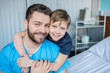 © LIGHTFIELD STUDIOS - Father and son hugging while sitting on hospital bed, dad and son in hospital