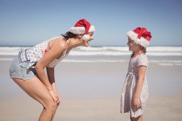 Wall Mural - Cheerful woman with daughter wearing Santa hat at beach