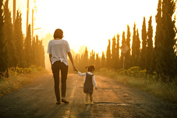 Wall Mural - Mother and little asian toddler girl walking together on the way at the sunset with copy space, view from the back