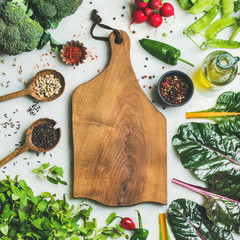 Fresh raw greens, unprocessed vegetables and grains over light grey marble kitchen countertop, wooden board in center, top view, copy space, square crop. Healthy, vegan, detox, dieting food concept