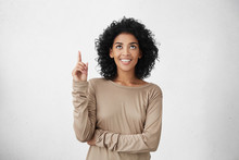 Waist Up Shot Of Joyful Girl Wearing Beige Long Sleeve T-shirt Looking Up, Pointing Finger At Copy Space Above Her Head. Black Young Woman Indicating Something On Blank Studio Wall With Hand