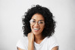 Positive human emotions. Portrait of beautiful and charming female student with Afro hairstyle, having shy look, laughing at camera, wearing stylish round eyeglasses, touching her neck with hand