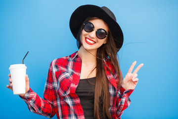 Wall Mural - Portrait of beauty fashion smiling woman with coffee, making peace by fingers in sunglasses on blue background. Outdoor