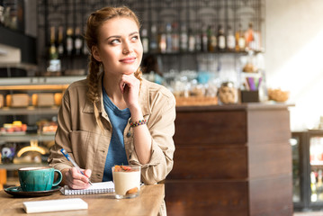 Pensive female writing into copybook in cafe