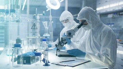 Wall Mural -  In a Secure High Level Laboratory Scientists in a Coverall Conducting a Research. Chemist Adjusts Samples in a  Petri Dish with Pincers and then Examines Them Under Microscope.