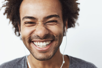 Wall Mural - Close up portrait of young happy african man smiling listening to upbeat streaming music laughing over white background. Youth concept.