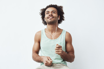 Wall Mural - Young african man listening to online streaming music in wired headphones dancing pretending to drum enjoying the bass over white background.