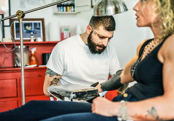 Wall Mural - Professional bearded tattooer preparing blond woman for special tattoo session inside ink trendy studio