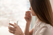 © fizkes - Young woman takes white round pill with glass of water in hand. Stressed female looking in window and drinking sedated antidepressant meds. Woman feels depressed, taking drugs. Medicines at work