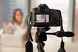 © fizkes - Close up image of camera on tripod with smiling woman on back screen and blurred scene on background. Recording video on modern DSLR camera. Personal videoblog social network concept