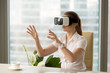 © fizkes - Businesswoman in VR headset interacting with virtual objects or interface while sitting in armchair at the table in office. Young girl making virtual business presentation with virtual reality glasses