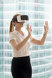© fizkes - Young woman in formal clothes enjoying virtual tour or excursion with VR headset. Girl in virtual reality glass standing near bright window and touching objects in virtual or augmented reality