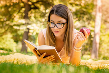 Outside Portrait Of Young Beautiful Woman With Apple Reading Book In Park