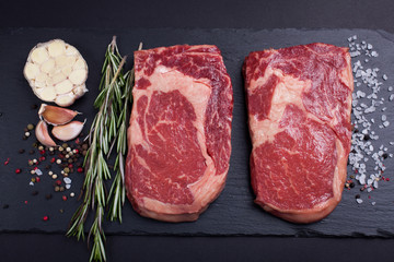 Two fresh raw marble meat, black Angus ribeye steak with spices on a dark stone background