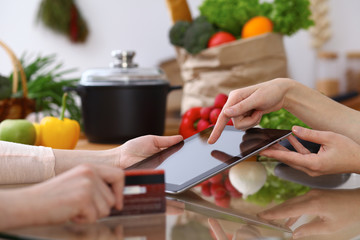 Human hands of two female persons using touchpad in the kitchen. Closeup of two women are making online shopping by tablet computer and credit card. Cooking and shopping concept.