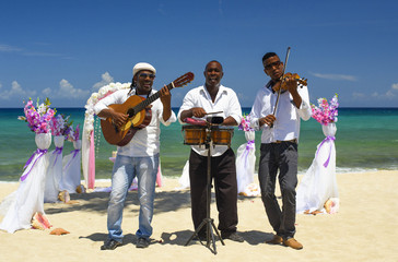 Fiddler, guitarist and drummer in white shirt and grey hat are playing musical instruments on the beach