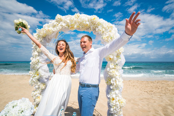 Beautiful wedding couple bride in lace wedding dress with bouquet of roses and groom reach their hands to sun near white arch on the beach of caribbean sea