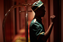 Side View Portrait Of African Bellboy Knocking On Hotel Room Door Delivering Luggage In Dim Hallway