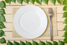 Empty Plate On A Bamboo Mat With A Fork, View From Top, On A Wooden Background, Framed With Leaves Of A Tree