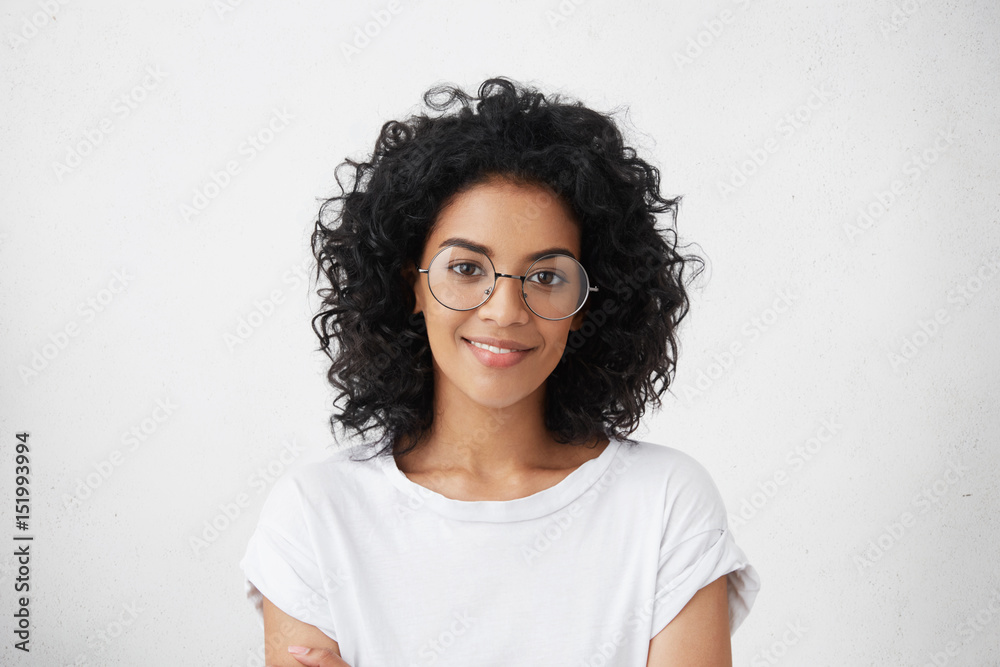Close up studio shot of beautiful young mixed race woman model with curly dark hair looking at camera with charming cute smile while posing against white blank copy space wall for your content - obrazy, fototapety, plakaty 